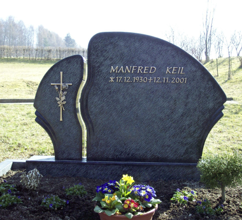Zweigeteilter Stein mit "Bruchstelle" dazwischen. Auf dem linken Teil ist ein Kreuz mit Blumen rechts steht der Name und die Lebensdaten